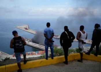 Men watch smoke rising from a dock after explosions were heard at La Guaira port, Venezuela, Saturday, Jan. 3, 2026. (AP Photo/Matias Delacroix)
