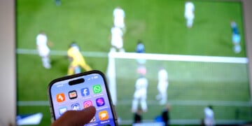 PRODUCTION - 05 June 2026, Baden-Württemberg, Stuttgart: A hand holds a cell phone while a soccer game is being played on a TV screen (staged scene) Photo: Bernd Weißbrod/dpa (Photo by Bernd Weißbrod/picture alliance via Getty Images)